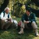 Two men sit on grass under trees, talking. One, an elderly man with white hair and a mustache, appears to be a Great Mentor. Both wear outdoor jackets; backpacks rest beside them in the dappled sunlight.