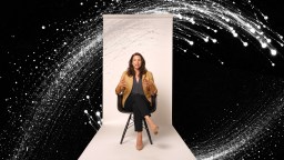 A woman sits on a chair against a white backdrop, gesturing with her hands, with a dynamic black background and white abstract swirl surrounding her.