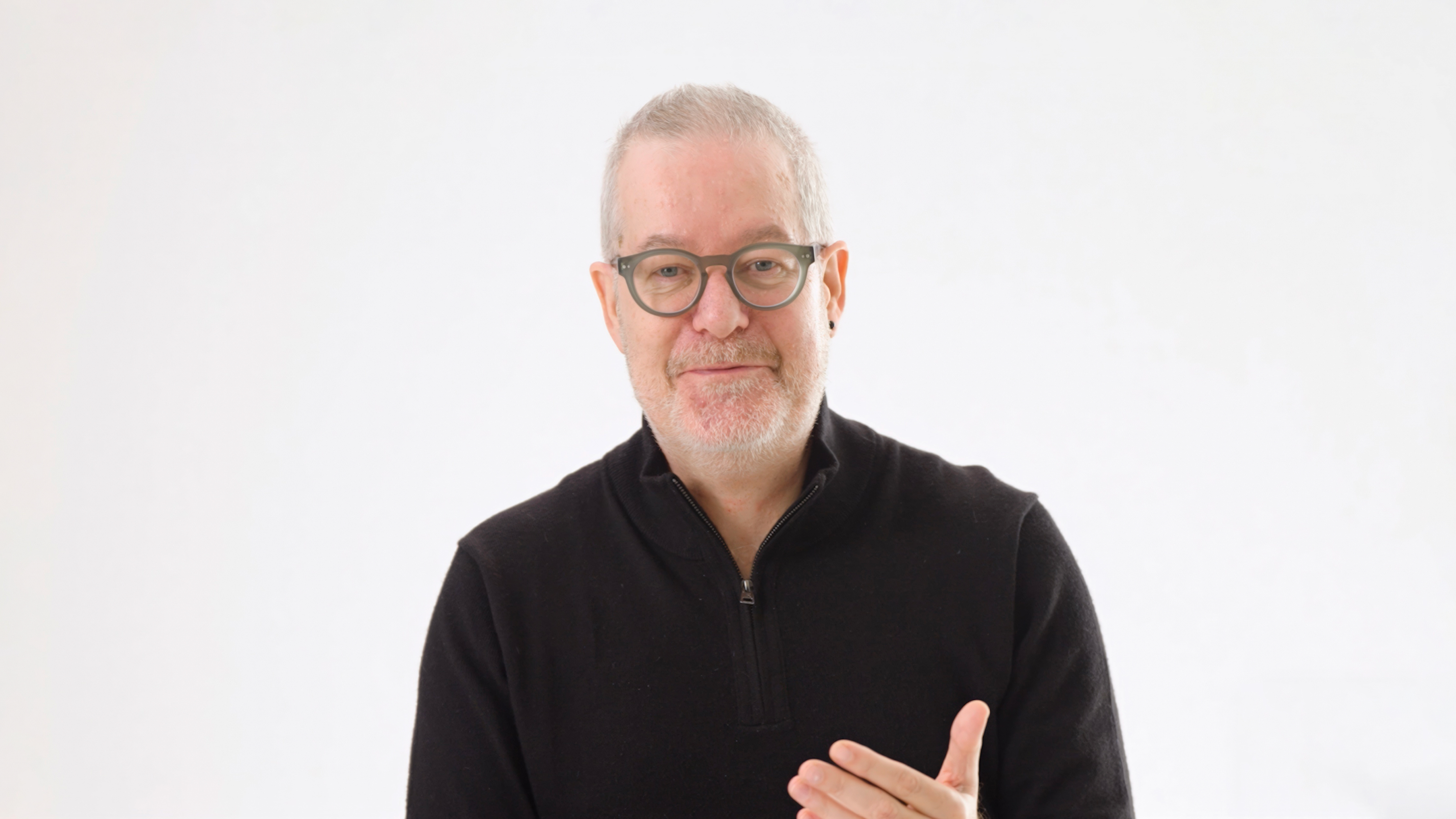 A middle-aged man with glasses and short gray hair, wearing a black pullover, stands against a plain white background, gesturing with his right hand.
