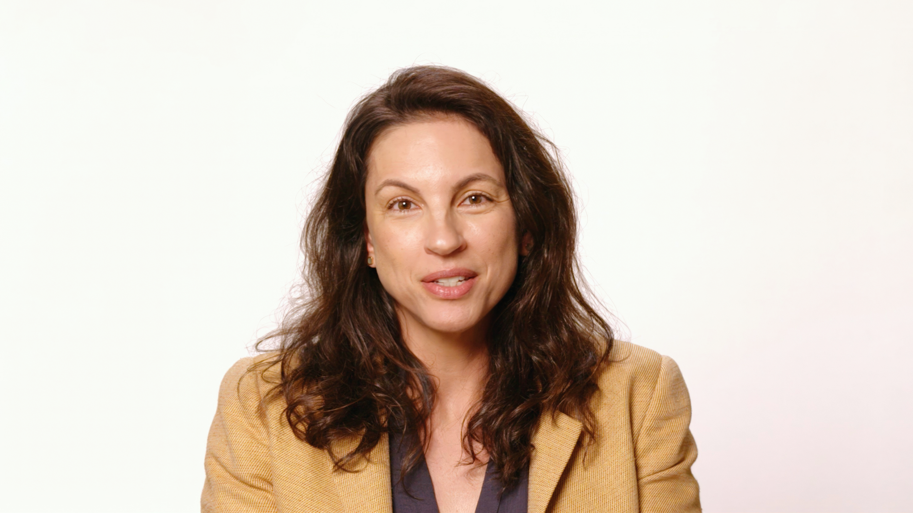 A woman with long brown hair wearing a tan blazer over a dark shirt sits in front of a plain white background, looking at the camera.