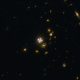 A deep space image shows numerous distant galaxies and stars against a dark background, including several bright spots shaped by a gravitational lens cross, with diffuse light sources scattered throughout.