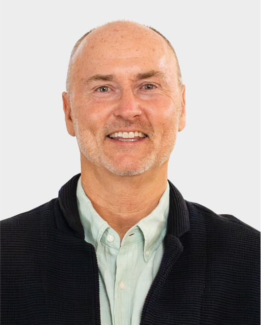 A middle-aged man with light skin, short thinning hair, and a light beard, wearing a light green shirt and a dark jacket, smiling at the camera against a plain background.