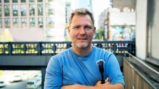Simon Squibb, a middle-aged man in a blue shirt, stands outdoors in an urban setting, holding a small microphone with his arms crossed.