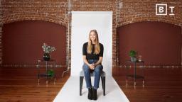 A woman sits on a chair in front of a white backdrop in a brick-walled room, with potted plants on tables on either side.